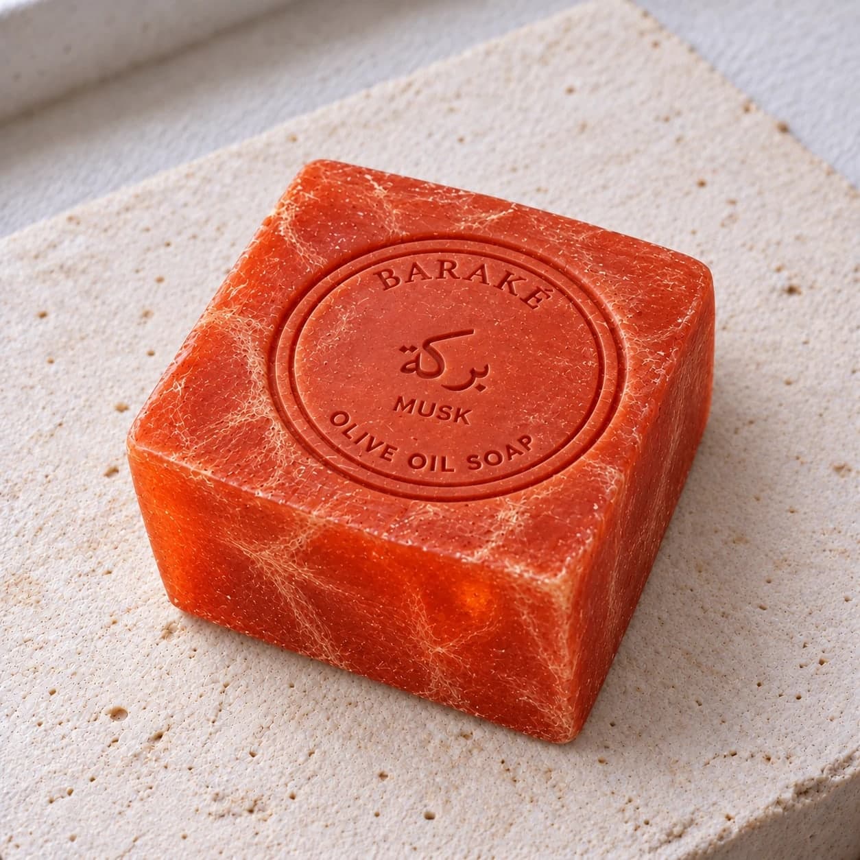 A square Baraké olive oil soap, musk, deep orange with pale veins running through it, the Baraké seal pressed into the top, on pale stone.