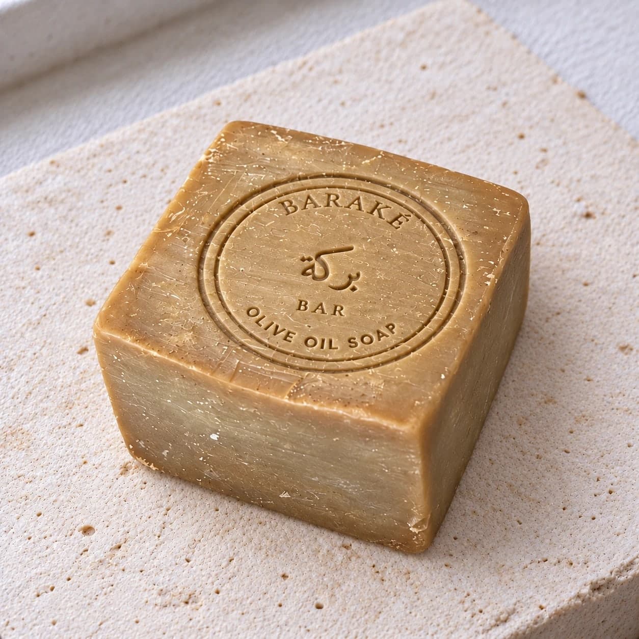 A square Baraké olive oil soap, original, warm golden olive with a rough hand-cut surface, the Baraké seal pressed into the top, on pale stone.